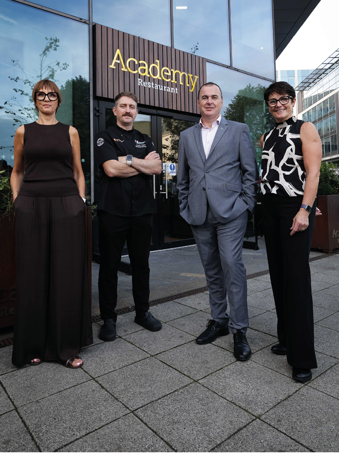 Four adults dressed in business and casual attire stand outside a building with a sign that reads Academy Restaurant. Description generated by AI