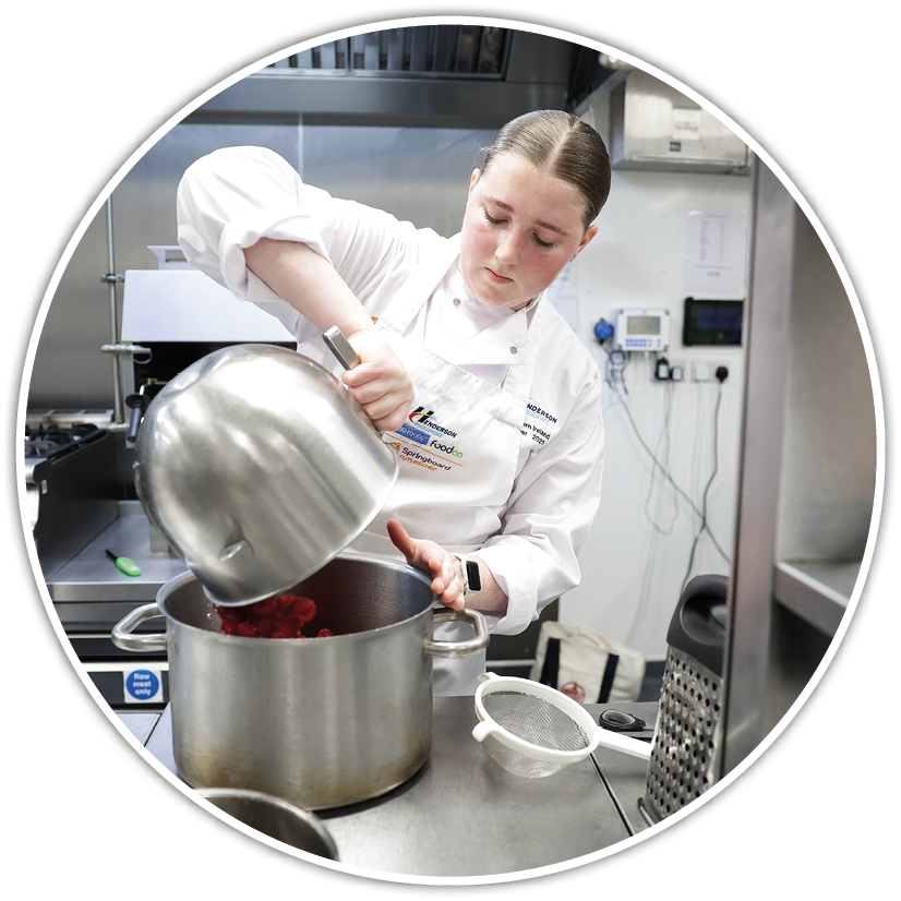 A person wearing a white chef's coat is pouring red ingredients from a large metal bowl into a pot in a professional kitchen. Description generated by AI