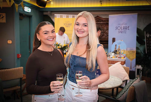 Two adults smiling and holding glasses of a beverage in a cozy indoor setting with banners and seating in the background. Description generated by AI