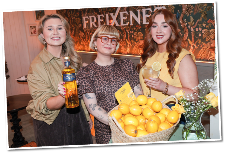 Three adults smiling and posing indoors with a basket full of lemons, one holding a bottle and another holding a glass with a lemon slice. Description generated by AI