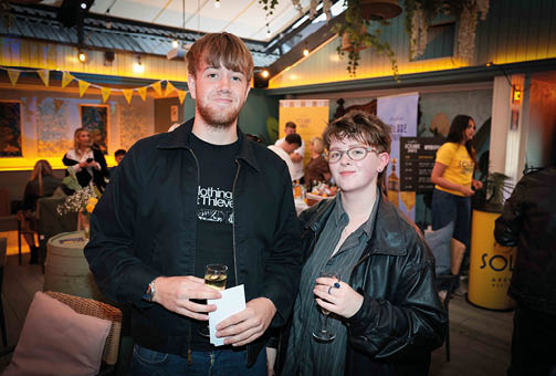 Two adults wearing casual jackets hold drinks and papers while standing in a warmly lit indoor space decorated with banners and plants, with other people and seating visible in the background. Description generated by AI