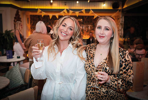 Two adults smiling and holding glasses of champagne in a warmly lit indoor setting with other people and festive decorations in the background. Description generated by AI