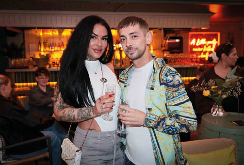 Two adults with long and short hair stand close together holding drinks in a warmly lit bar with other people seated and socializing in the background. Description generated by AI