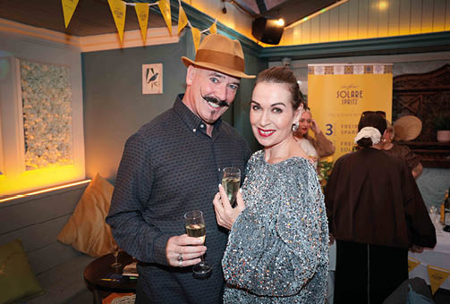 Two adults dressed in stylish clothing are smiling and holding glasses of a sparkling beverage at a festive indoor gathering. Description generated by AI