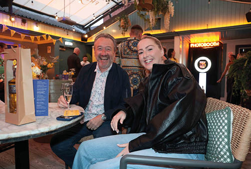 Two adults smiling and sitting at a table in a warmly lit indoor setting with one holding a glass of champagne and a photo booth visible in the background. Description generated by AI