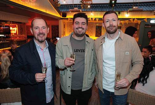 Three adults smiling and holding glasses of champagne in a warmly lit indoor setting with other people in the background. Description generated by AI