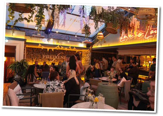 A lively indoor restaurant or bar scene with people sitting and standing around tables under hanging plants and warm lighting, featuring a neon sign and a cozy atmosphere. Description generated by AI