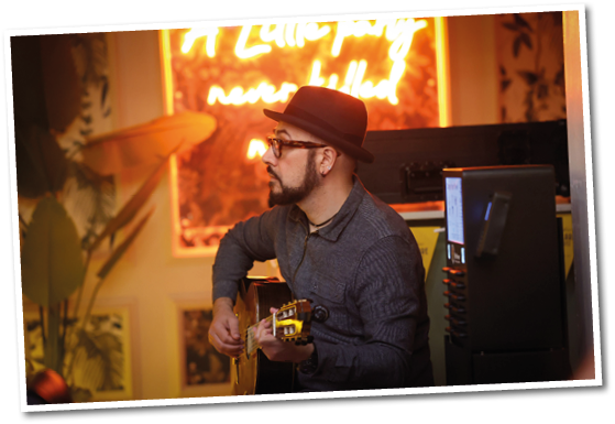 A person wearing a hat and glasses is playing an acoustic guitar in a warmly lit room with a neon sign and plants in the background. Description generated by AI