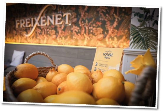 A basket filled with lemons is placed in front of a decorative wall with the word Freixenet and a sign promoting Solare Spritz. Description generated by AI
