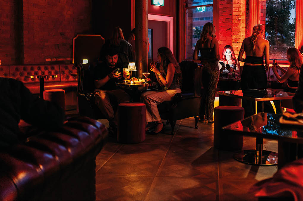 People sit and stand in a dimly lit lounge with warm red lighting, engaging in conversation and using their phones. Description generated by AI