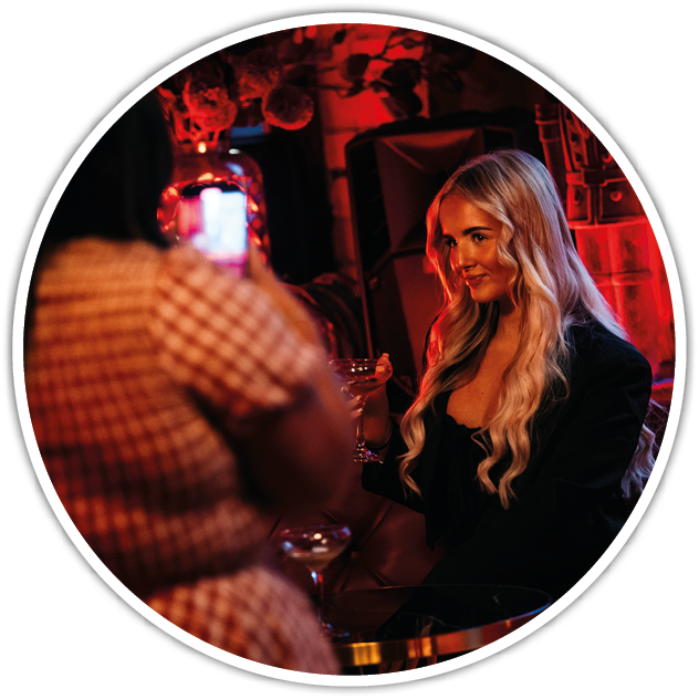 Two adults are in a dimly lit setting with red lighting, one holding a drink and smiling while the other takes a photo with a smartphone. Description generated by AI