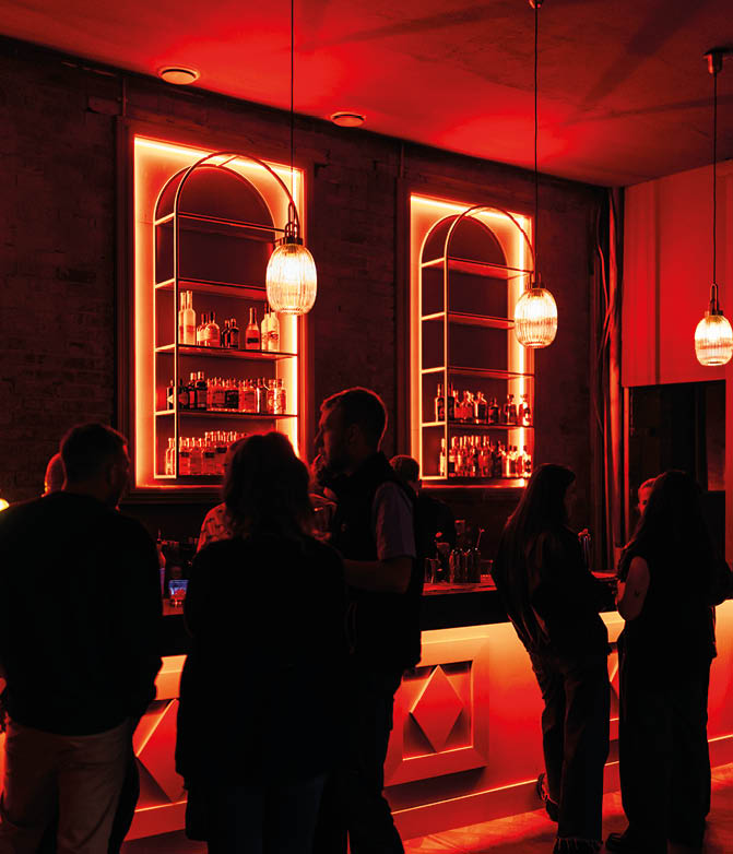 People are gathered and conversing at a bar illuminated by warm red neon lights and hanging pendant lamps. Description generated by AI
