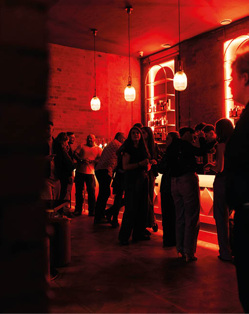 People are socializing and standing near a bar with warm red lighting and hanging pendant lamps in a cozy indoor setting. Description generated by AI
