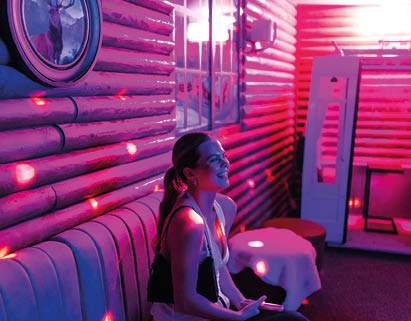 A person with long hair tied back is sitting on a cushioned bench in a dimly lit room with purple and pink lighting and a framed deer picture on the wall. Description generated by AI