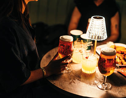 Two people sit at a dimly lit round wooden table with glasses of beer, cocktails, and a small lamp in the center. Description generated by AI