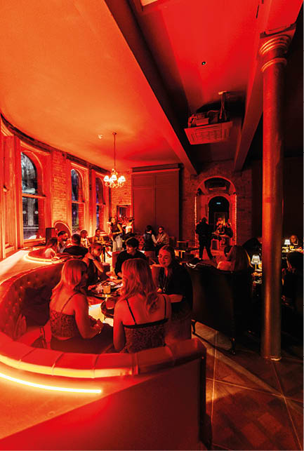 People dressed in stylish clothing are seated and socializing in a warmly lit, modern lounge with large windows and exposed brick walls. Description generated by AI