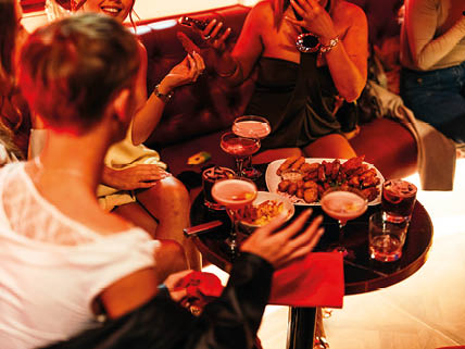 People dressed in stylish clothing are sitting around a small round table with drinks and a plate of appetizers in a warmly lit social setting. Description generated by AI