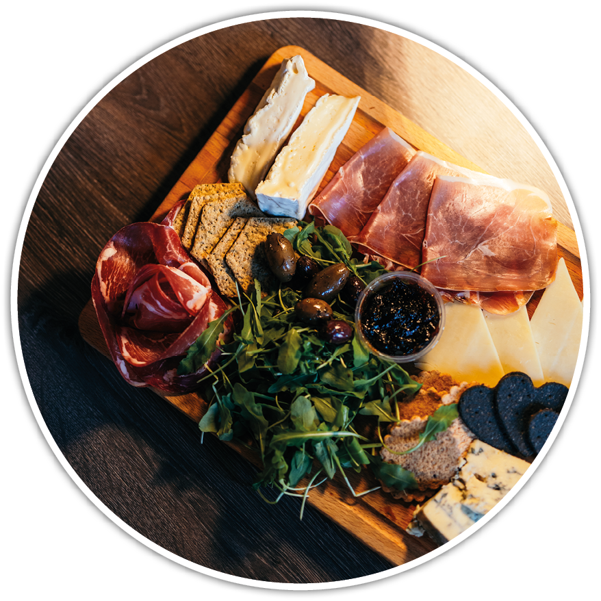 A wooden board on a dark surface holds an assortment of sliced cured meats, cheeses, crackers, olives, greens, and a small container of dark spread. Description generated by AI