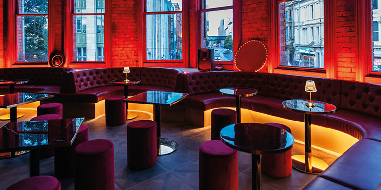 A modern lounge area features curved maroon seating with round black tables and soft orange lighting under large windows showing an urban street view. Description generated by AI