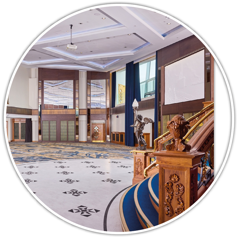 A spacious, elegant interior features a patterned floor, ornate wooden staircase with blue carpet, and a bronze statue holding a torch near large windows and high ceilings. Description generated by AI