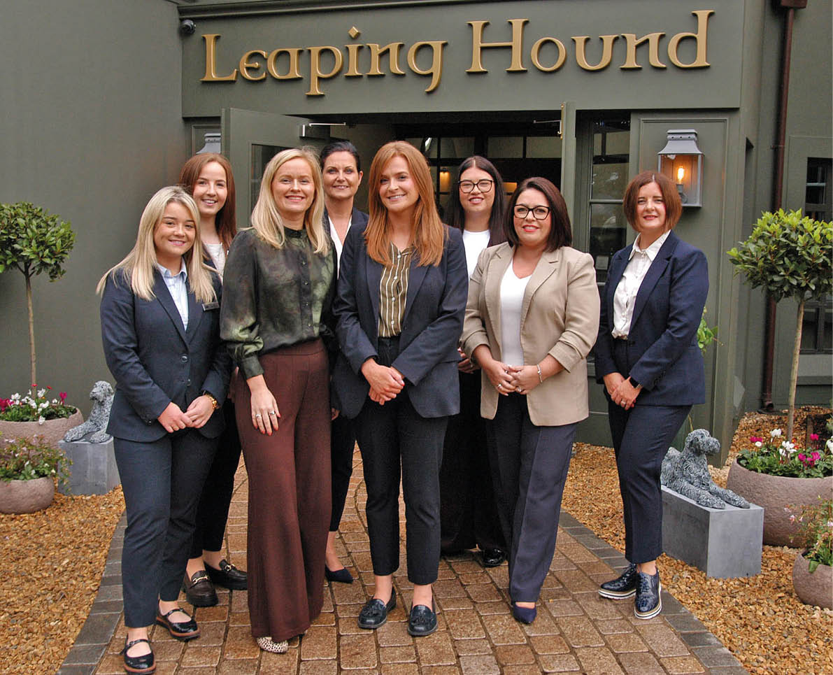 Seven adults dressed in business casual attire stand together smiling outside a building with a sign that reads Leaping Hound. Description generated by AI