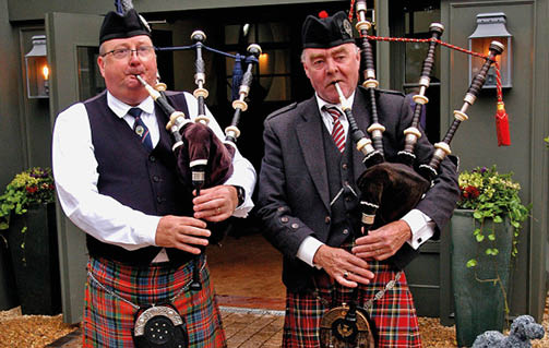 Two adults wearing traditional Scottish attire including kilts and hats are playing bagpipes outside a building decorated with plants. Description generated by AI