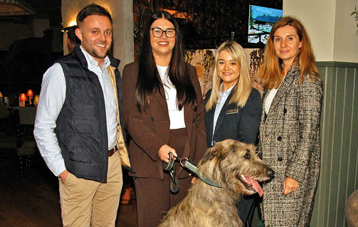 Four adults dressed in business casual attire stand indoors smiling with a large dog sitting in front of them. Description generated by AI