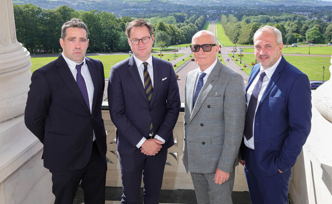 Four adults dressed in formal suits stand side by side outdoors on a balcony with a scenic view of a tree-lined pathway and distant landscape behind them. Description generated by AI