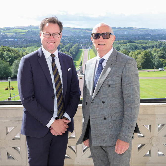 Two adults dressed in suits stand side by side outdoors with a scenic view of a long tree-lined pathway and green landscape in the background. Description generated by AI