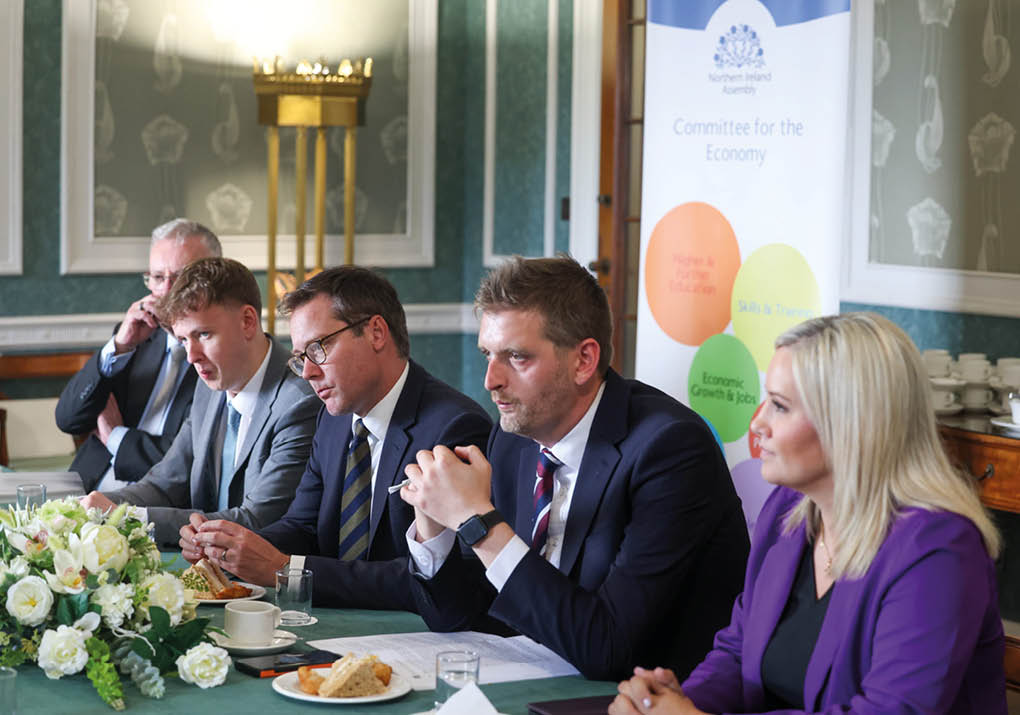 Five adults in formal attire are seated at a table with documents, food, and drinks, engaged in a meeting in a decorated room with a banner labeled Committee for the Economy in the background. Description generated by AI