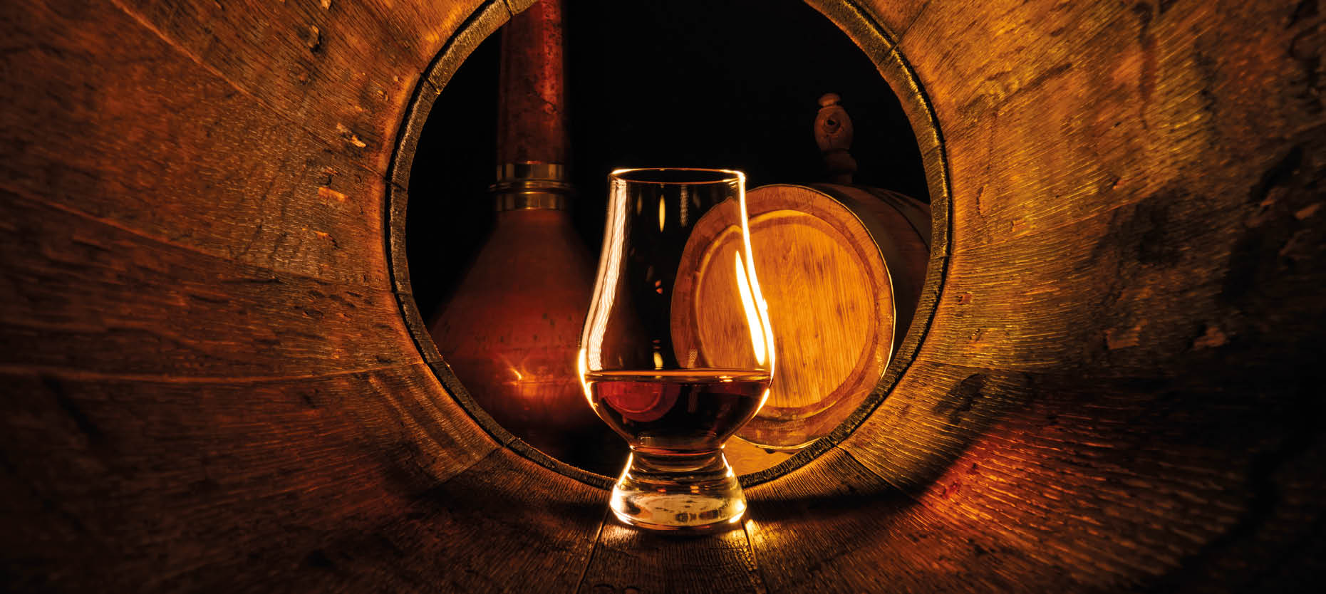 A glass of whiskey is placed inside a wooden barrel with a copper still and another barrel visible in the background. Description generated by AI