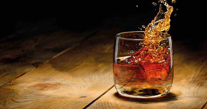 A glass of whiskey with ice is shown on a wooden surface with liquid splashing out of the glass against a dark background. Description generated by AI