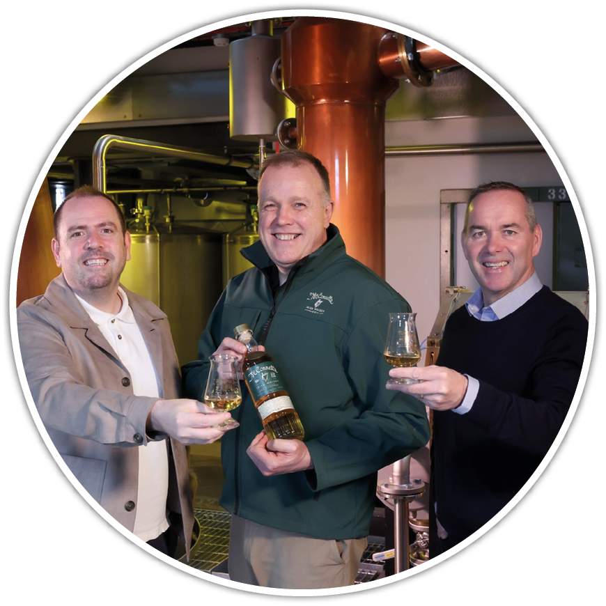 Three adults smiling and holding glasses of whiskey with one person also holding a bottle in an industrial setting. Description generated by AI
