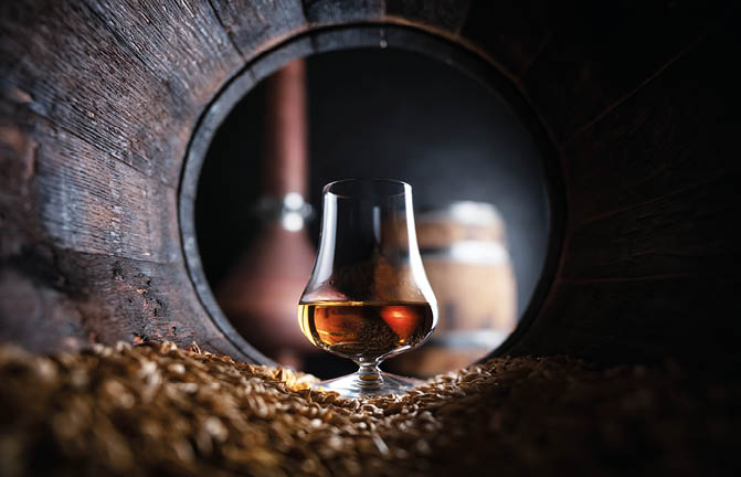 A glass of amber liquid is placed inside a wooden barrel surrounded by grains with another barrel blurred in the background. Description generated by AI