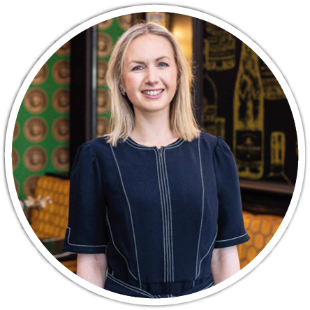 A person with shoulder-length blonde hair is smiling and wearing a dark dress with white stitching, standing indoors with patterned green and yellow seating in the background. Description generated by AI