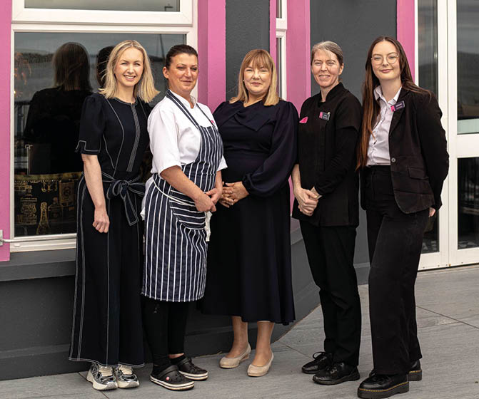 Five adults stand side by side outside a building, with one wearing a striped apron and others dressed in dark professional or casual attire, smiling and posing for the photo. Description generated by AI