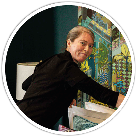 A person with tied-back hair wearing a black long-sleeve shirt is reaching toward a colorful patterned wall in a room with a lamp and cushions. Description generated by AI