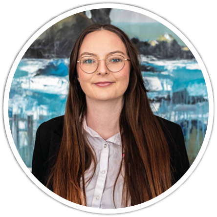 Person with long straight hair and glasses wearing a white button-up shirt and black blazer standing in front of a blue and white abstract painting. Description generated by AI