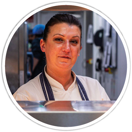 A person wearing a white chef's coat and apron stands behind a counter in a kitchen setting with a focused expression. Description generated by AI