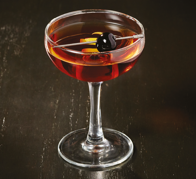 A cocktail glass filled with a dark amber drink garnished with a black cherry and a twist of citrus peel on a dark wooden surface. Description generated by AI