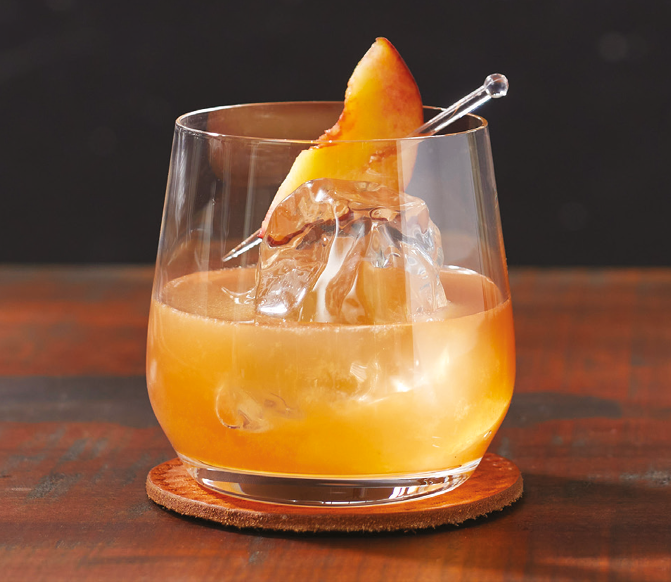 A glass with an orange-yellow cocktail on a wooden coaster contains a large ice cube and is garnished with a peach slice on a clear pick. Description generated by AI