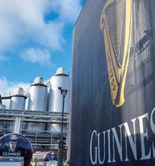 Large silver brewing tanks are visible behind a tall black banner featuring a golden harp and the word Guinness against a partly cloudy blue sky. Description generated by AI