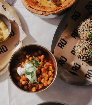 A bowl of chickpea stew topped with a dollop of cream and fresh herbs is placed on a table next to a plate with sesame-coated balls and a wooden bowl with flatbread. Description generated by AI