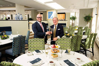 Two adults in suits are smiling and clinking glasses while standing behind a table set with teacups, plates, and a tiered tray of pastries in a bright, elegantly decorated dining room. Description generated by AI