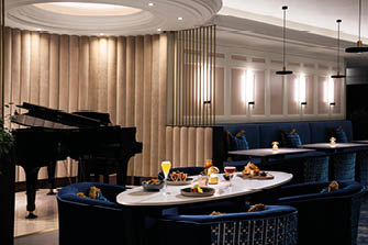 A modern lounge area features a grand piano, a white oval table set with drinks and food, surrounded by blue upholstered chairs and a blue cushioned bench against a wall with soft lighting. Description generated by AI