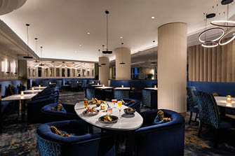 A modern, elegant restaurant interior features blue upholstered chairs and sofas around white tables with plates of food and drinks, illuminated by warm pendant and recessed lighting. Description generated by AI