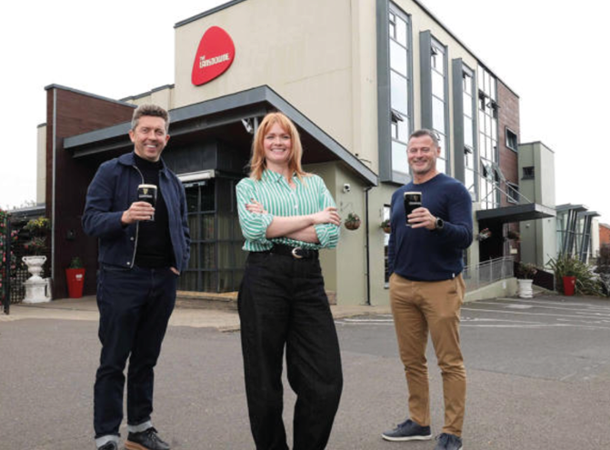 Three adults stand outside a modern building holding glasses of dark beer, with one person in a striped shirt and black pants smiling confidently in the center. Description generated by AI
