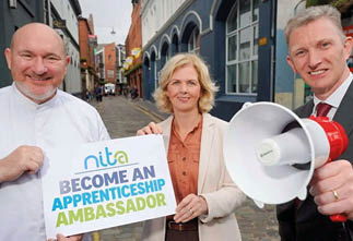 Three adults stand on a cobblestone street with one holding a sign that says “Become an Apprenticeship Ambassador" and another holding a megaphone. Description generated by AI
