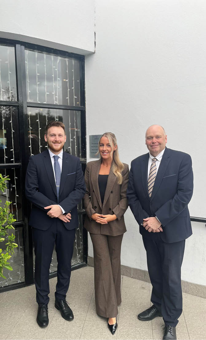 Three adults dressed in formal business attire stand side by side smiling in front of a building with glass doors and a white wall. Description generated by AI