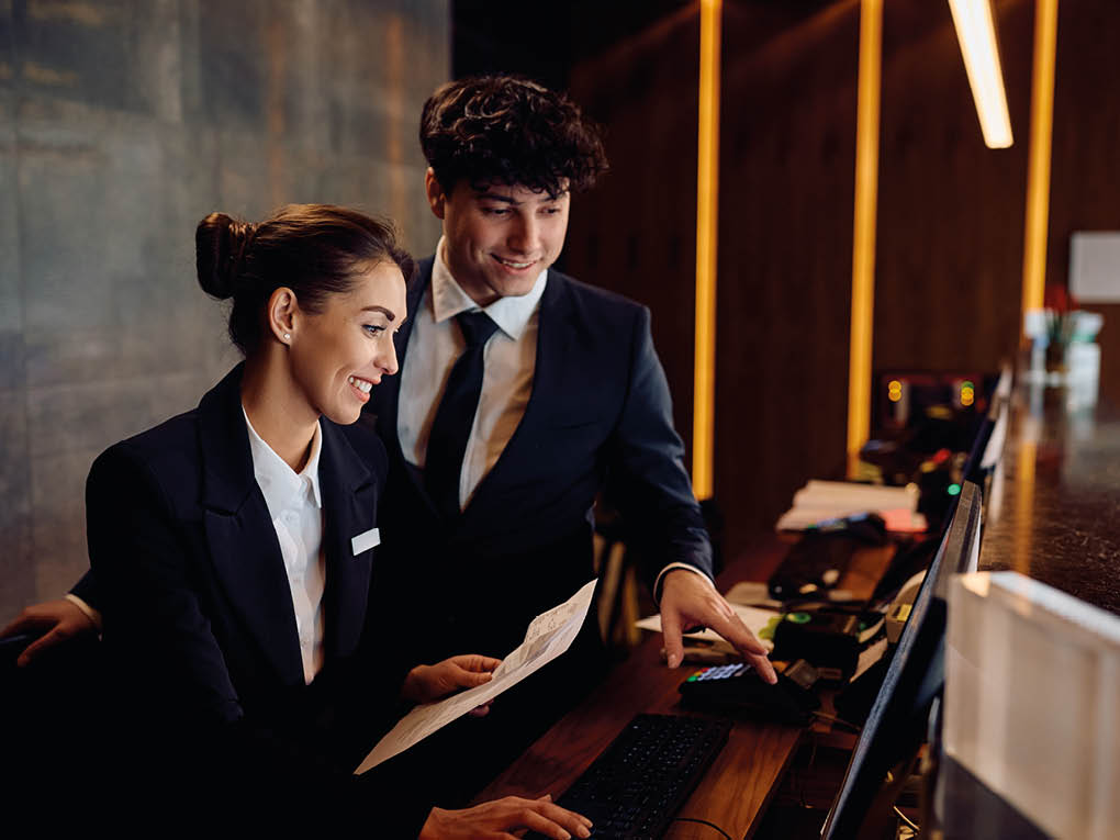 Two adults dressed in formal business attire are smiling and working together at a desk with a computer and documents in a modern office setting. Description generated by AI